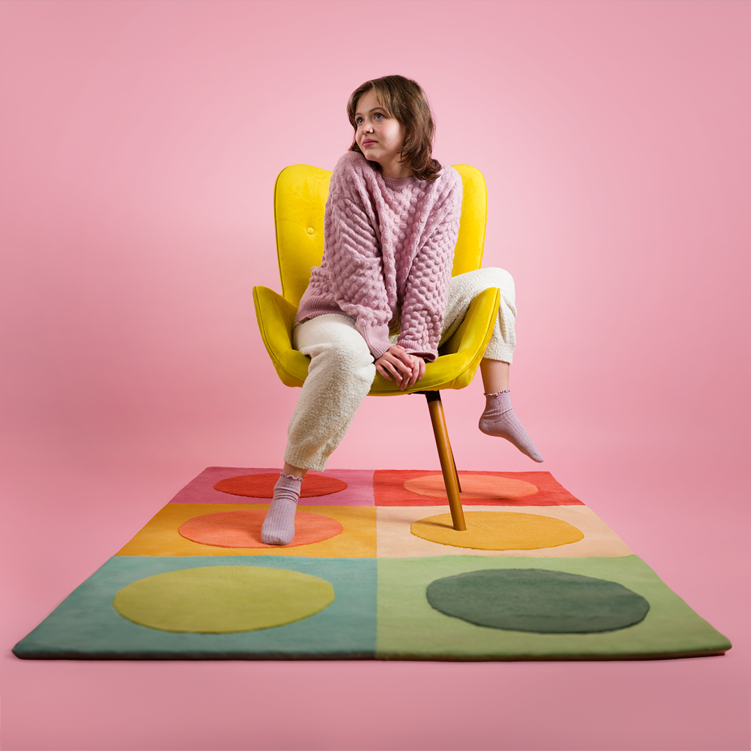 Woman sitting on a yellow chair on top of a polka dot rug that is 4 feet by 6 feet wide. There are two rows of squares with a polka dot in each square. Each polka dot and square is a different color.