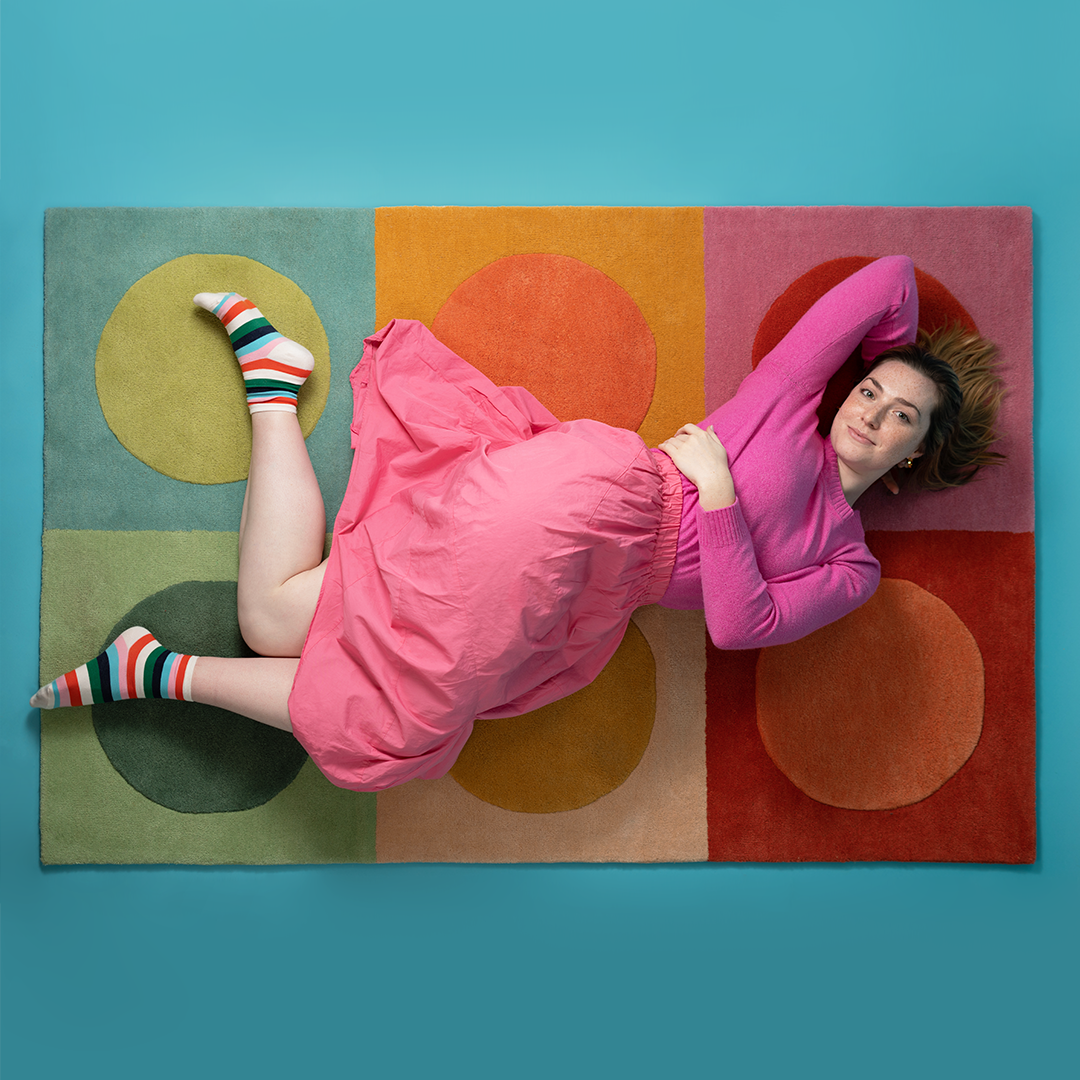 Woman laying on top of a polka dot rug that is 4 feet by 6 feet wide. There are two rows of squares with a polka dot in each square. Each polka dot and square is a different color.