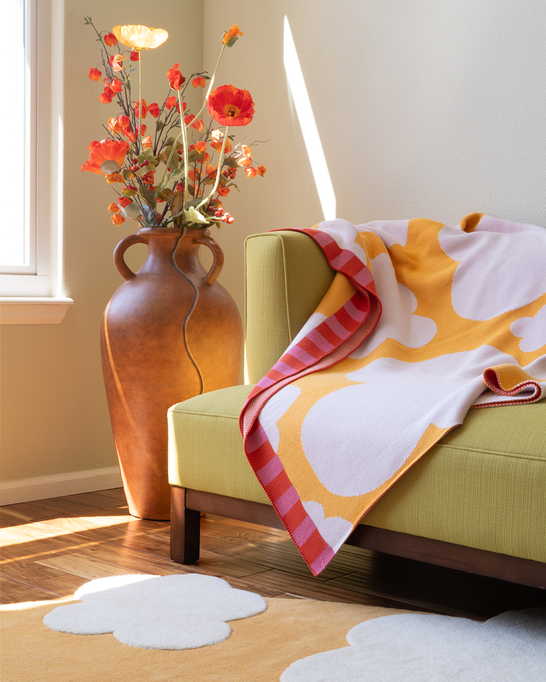 Yellow blanket with beige clouds and a border that is striped pink and orange. The blanket is sitting on a couch in a living room with a yellow cloud rug on the floor and a large vase with flowers on the floor.