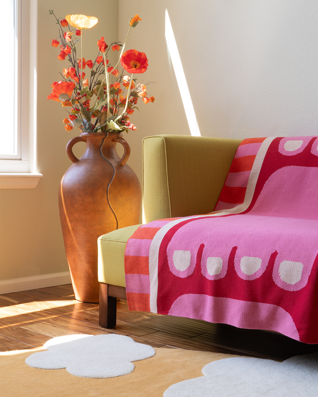 Photo of a living room with a pink and red blanket laying on the edge of a green couch. The pattern on the blanket looks like a pink foot on a red background with a striped border that is pink and orange.