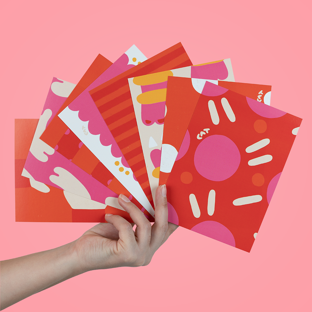 Hand holding eight pink and red postcards with a variety of shades and patterns on them. The first postcard is polka dotted and other variations include ruffly stripes, clouds, and noodle shapes.