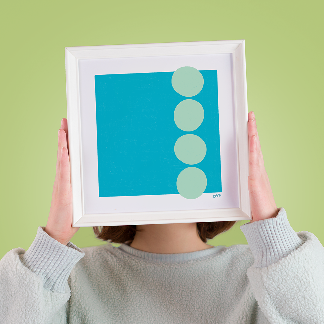 Photo of a woman holding a frame to her face with a print of a darker blue square and four light blue polka dots lined up vertically on the right side.
