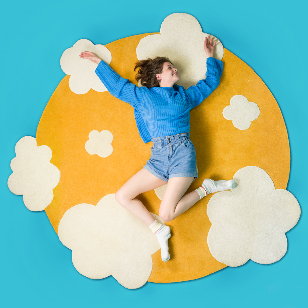 Woman in a blue sweater and jean shorts laying on a yellow rug with white clouds.