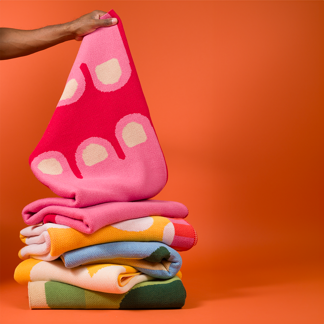 Hand holding up a pink and red blanket on a pile of other colorful blankets. 