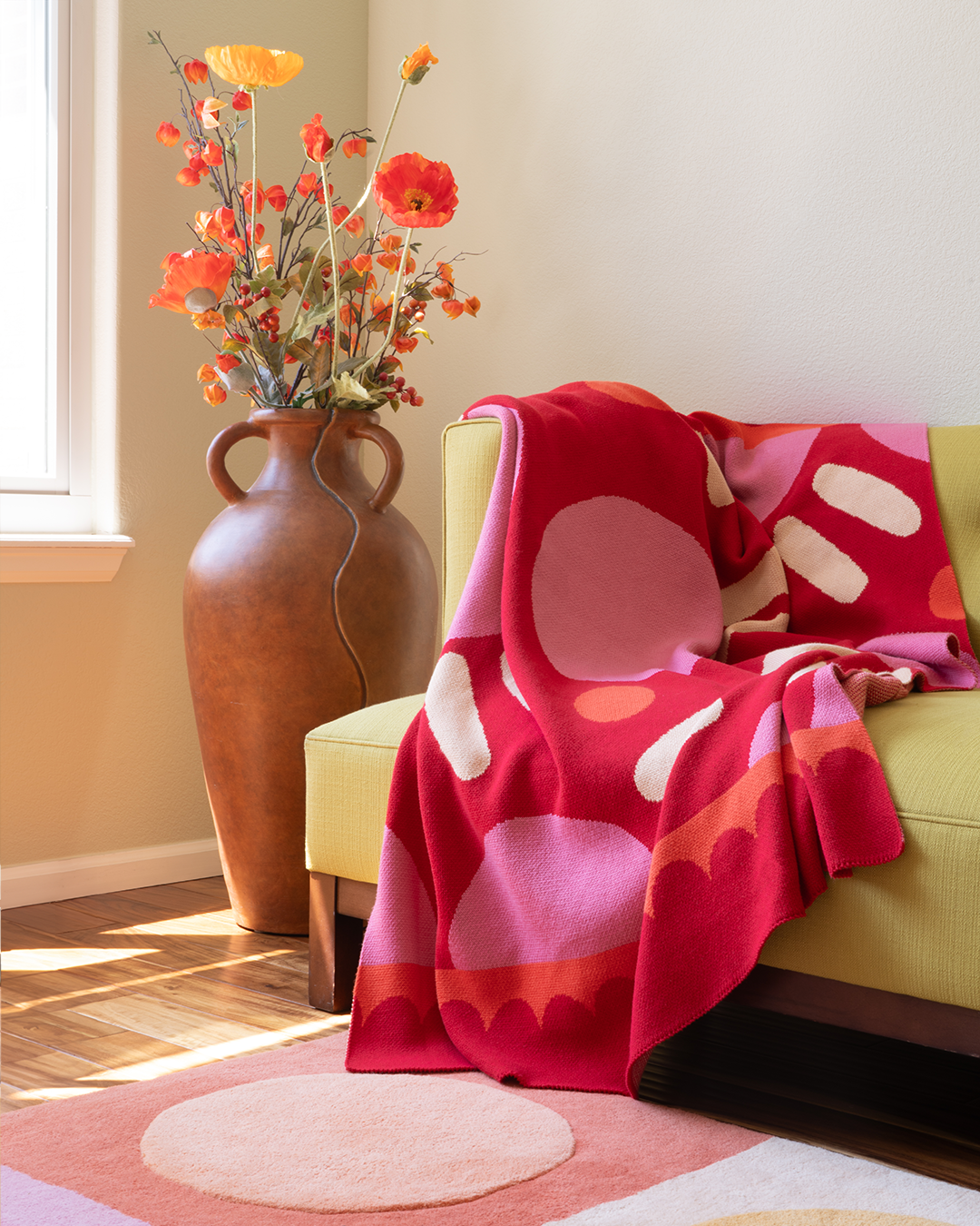 Photo of a living room couch with a red and pink polka dotted blanket splaying out of the corner of the couch. There is also a polka dotted rug on the floor and a giant vase of flowers sitting on the corner.