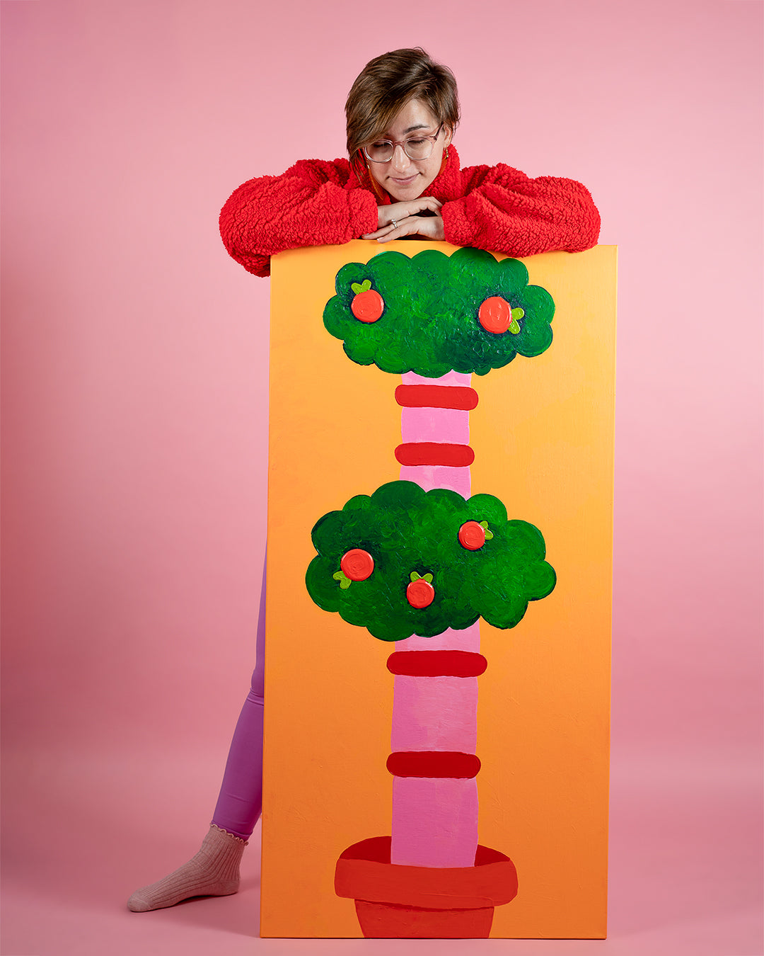 Photo of Cat Schmitz standing behind a four foot tall painting of a bushy tree with green leaves, oranges, and a pink and red striped tree trunk sitting in a red pot.