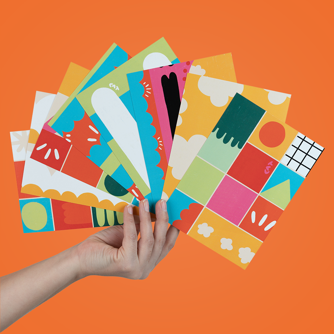 Hand holding eight multicolored postcards with a variety of shades and patterns on them. The first postcard is polka dotted and other variations include grid patterns, clouds, and noodle shapes.