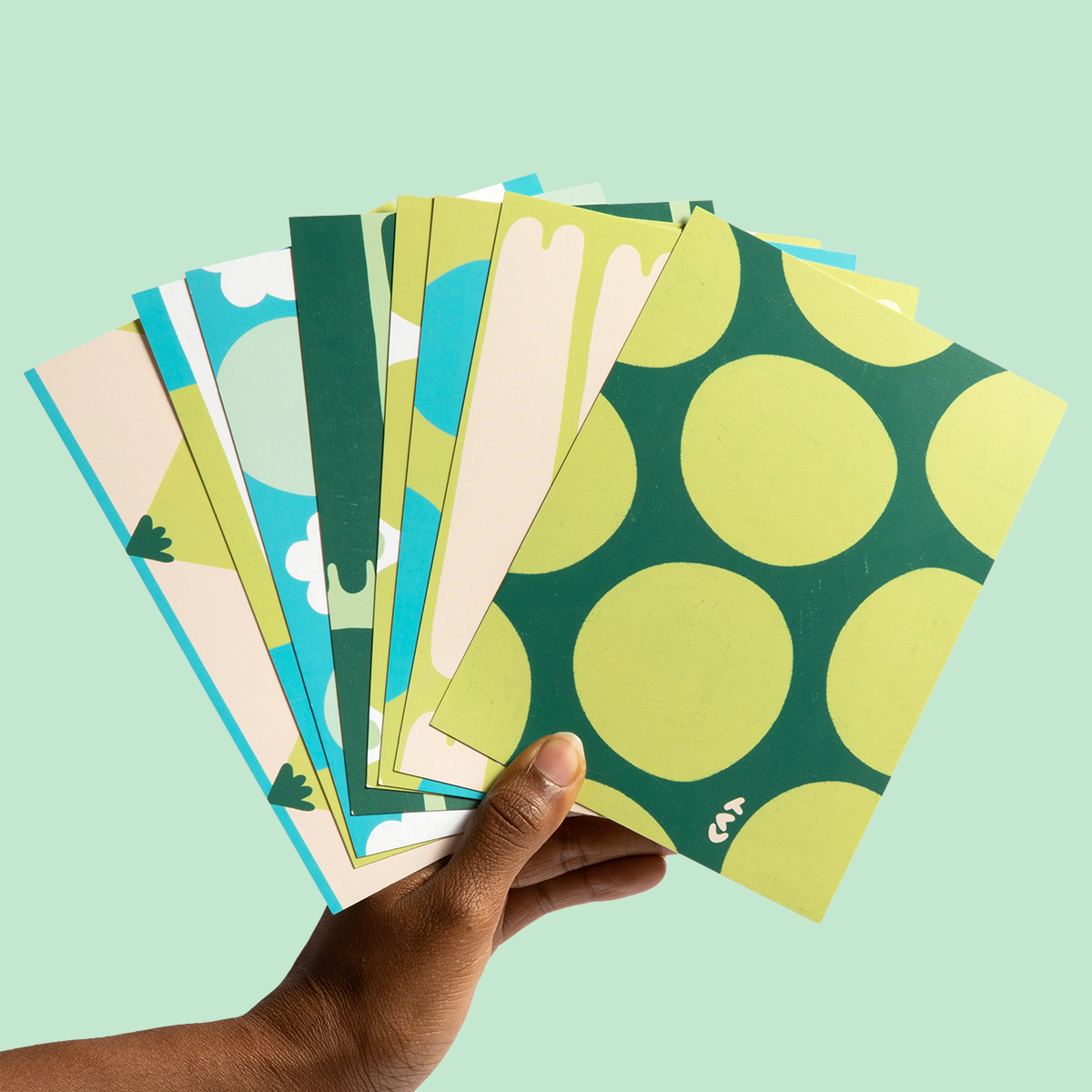 Hand holding eight green postcards with a variety of shades and patterns on them. The first postcard is polka dotted and other variations include mountains, clouds, and noodle shapes.